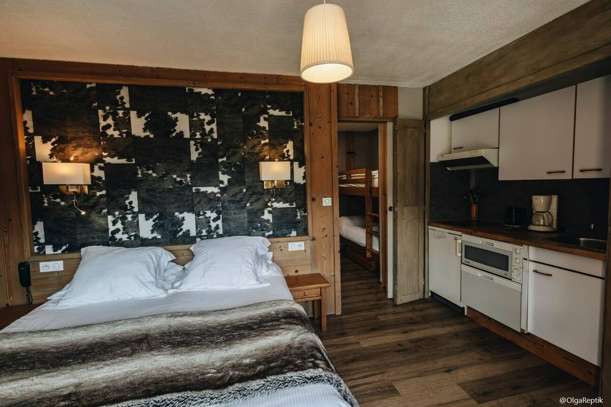 A bedroom with a large bed, wooden headboard, and bedside lamps. There is a kitchenette with white cabinets and a stove.