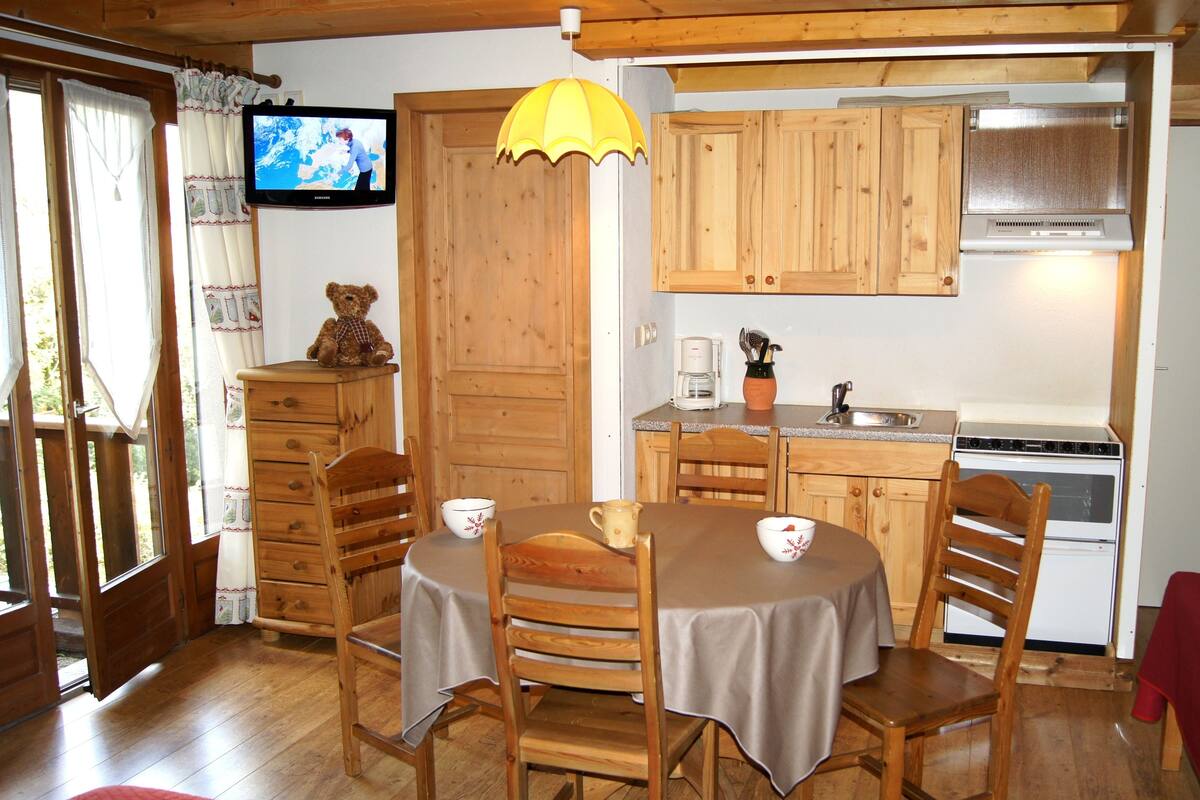 A cozy room with wooden furniture, a dining table, chairs, a kitchen area with a stove, and a television on the wall.