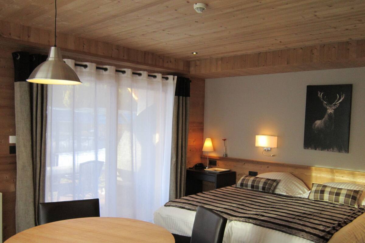 A hotel room with a wooden ceiling, a bed with a striped blanket, a bedside table with a lamp, a wall-mounted picture of a deer, and a round wooden table with chairs.