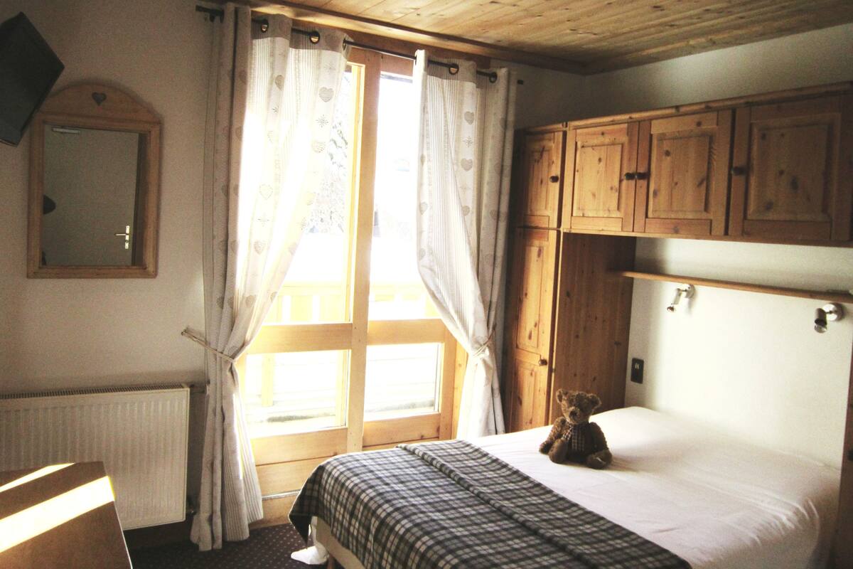 A bedroom with a wooden ceiling, a bed with a checkered bedspread, a teddy bear on the bed, and wooden cabinets above the bed.