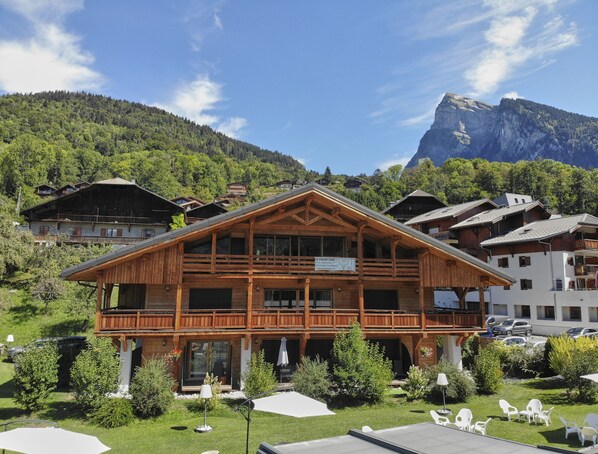 A wooden chalet with a balcony, surrounded by greenery and a mountain backdrop.