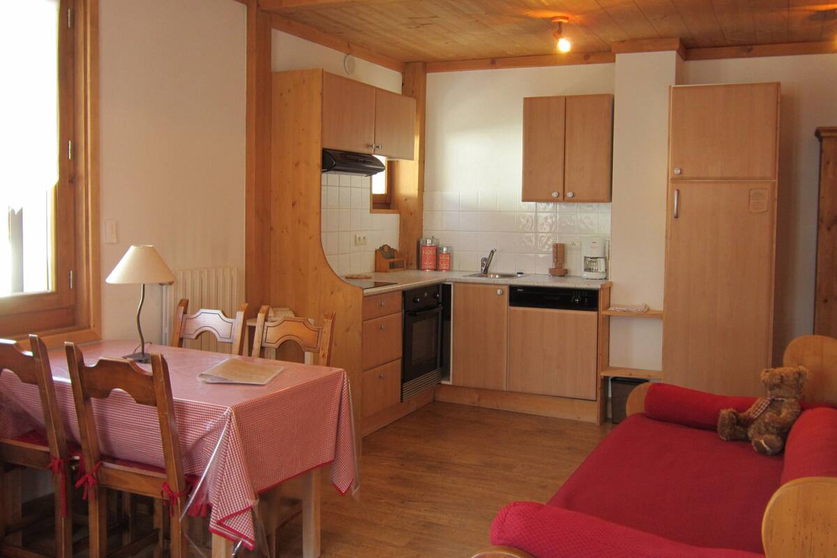 A compact kitchen with wooden cabinets, a dining area with a table and chairs, and a red sofa with a teddy bear.