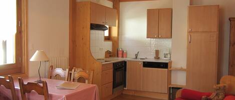 A compact kitchen with wooden cabinets, a dining area with a table and chairs, and a red sofa with a teddy bear.