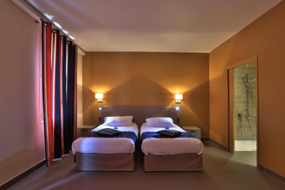 A hotel room with two beds, nightstands, wall sconces, a shower area, and a curtain.