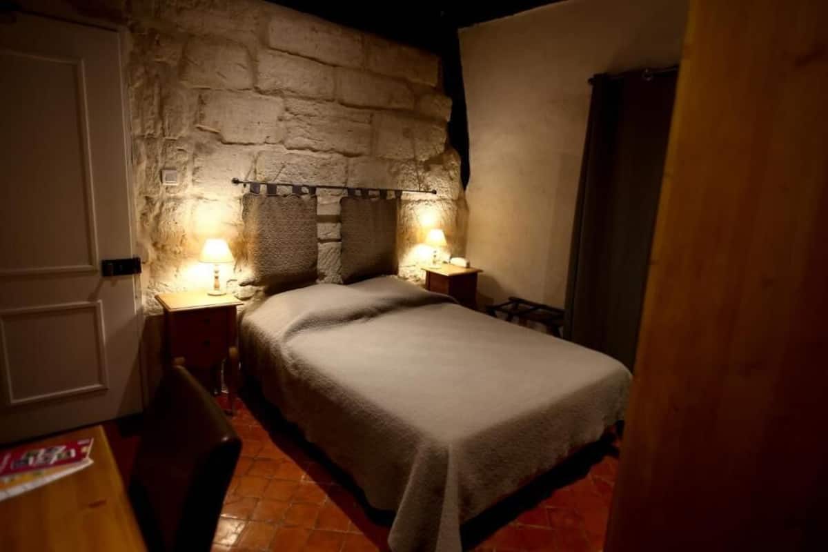 A bedroom with a stone wall, a bed with a blanket, two bedside tables with lamps, and a wooden door.
