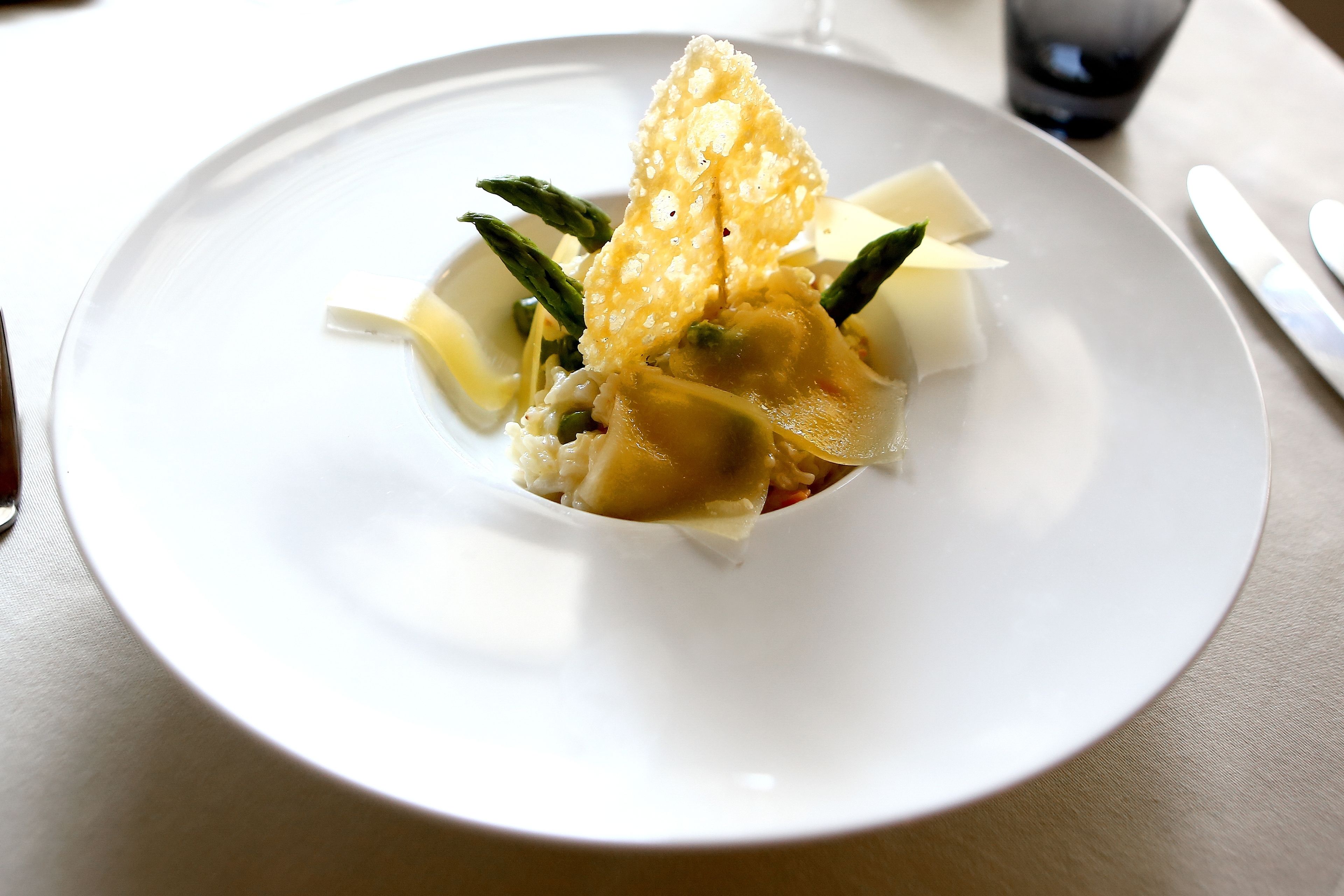 A white plate with a dish that includes asparagus and a crispy element, possibly a cracker or pastry, on a table with a knife and a glass of water.