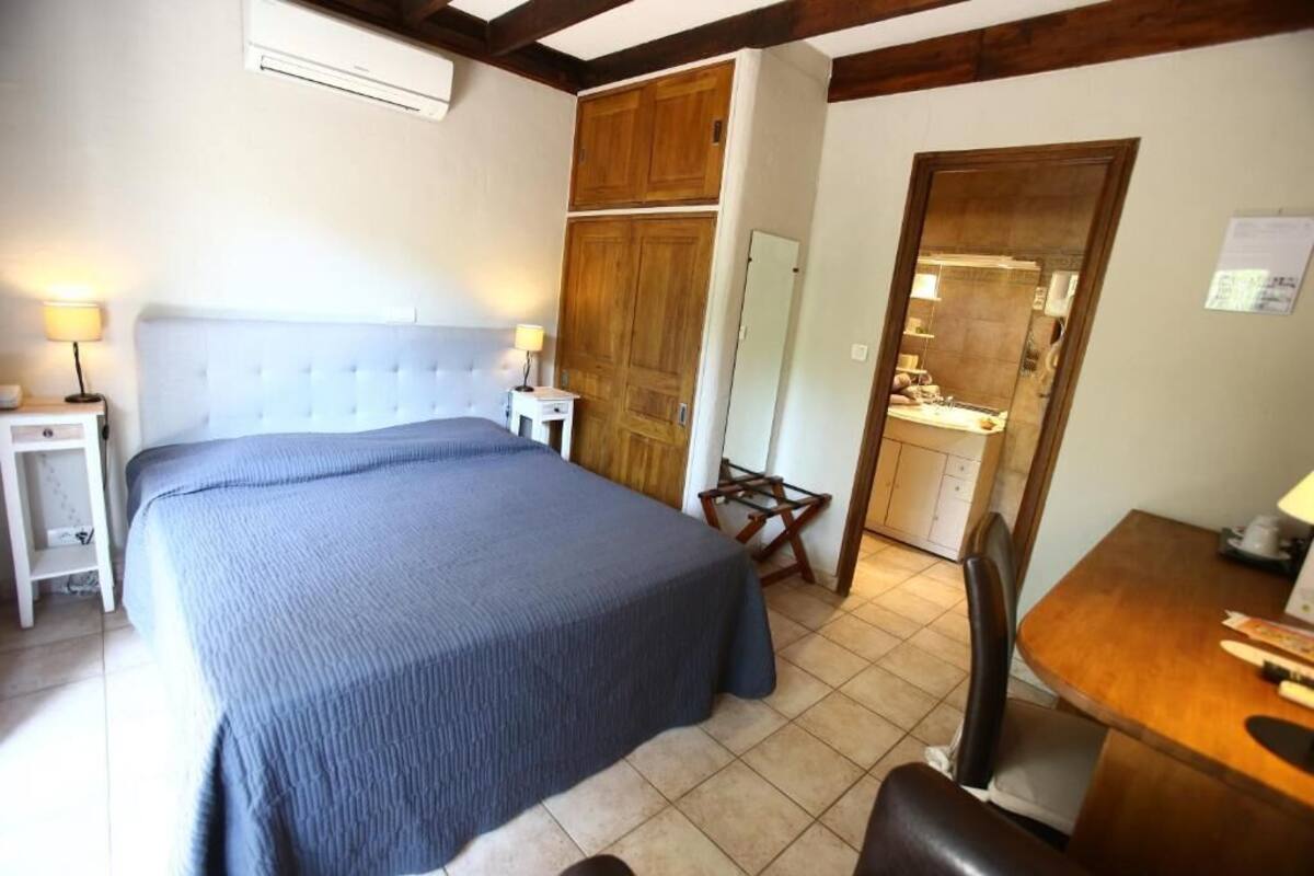 A bedroom with a bed, bedside tables, a wooden wardrobe, a small dining table, and a kitchen area.