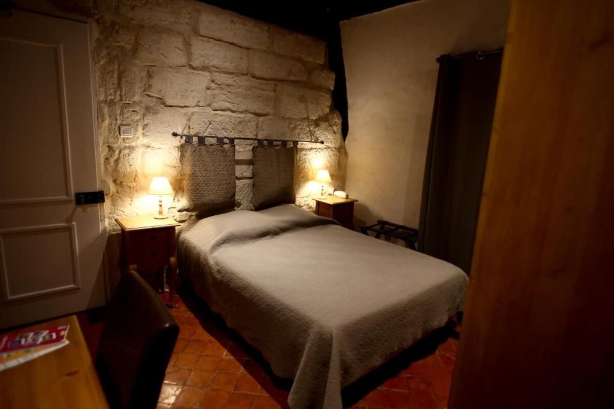 Un dormitorio con pared de piedra, una cama con manta, dos mesitas de noche con lámparas y una puerta de madera.