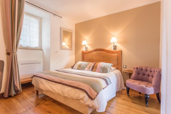 A bedroom with a wooden headboard, a bed with striped bedding, two wall-mounted lamps, a chair, and a window with curtains. - Le Troubadour (Rocamadour)