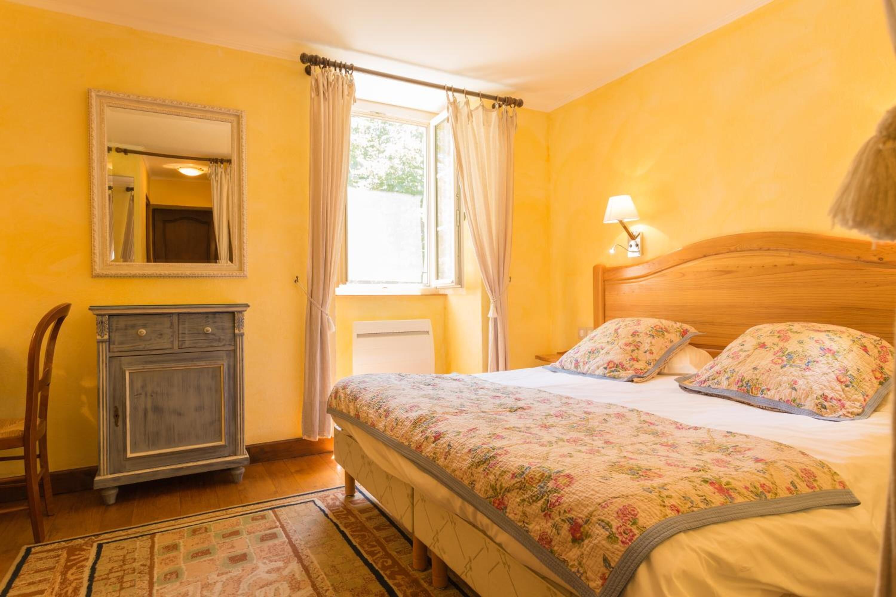 Une chambre à coucher avec un lit, une table de chevet, un miroir, une fenêtre avec des rideaux et une lampe.