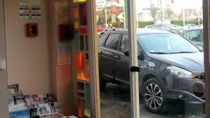 A car parked outside a building with a glass door, a small display of items inside, and flags in the background. - Hôtel Restaurant Le Hussard (Alencon)