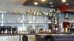 A bar area with shelves stocked with bottles and glasses, a counter, and a seating area with chairs. - Hôtel Restaurant Le Hussard (Alencon)