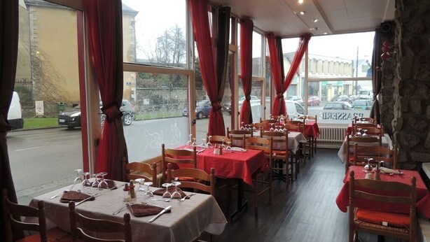 A restaurant with tables set for dining, large windows with red curtains, and a view of the street outside. - Hôtel Restaurant Le Hussard (Alencon)