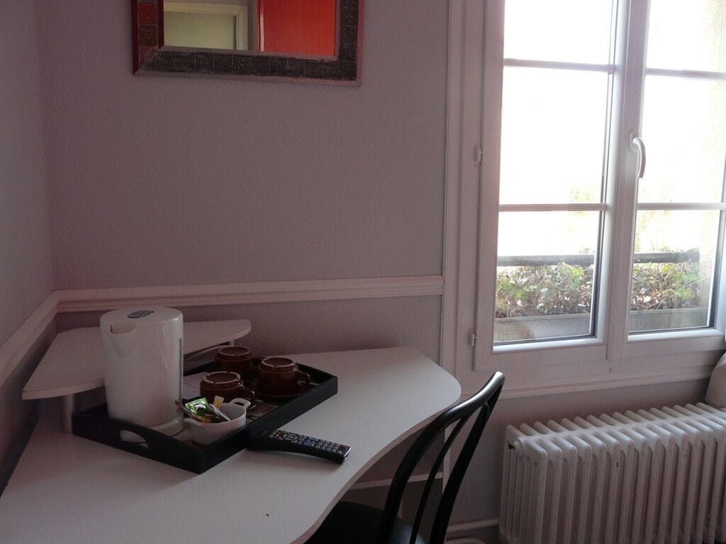 A hotel room with a white desk, a remote control, a tray with cups and a tissue box, and a window with a view of greenery outside.