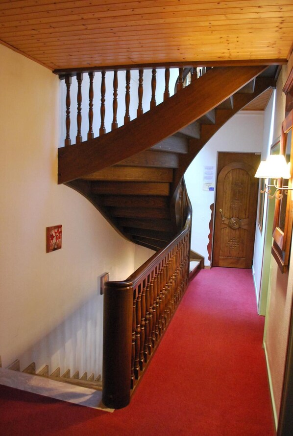 A wooden staircase with a red carpeted landing area, a wooden door, and a wall-mounted light fixture. - Kleiber (Saint-Jean-Saverne)