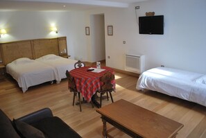 Une chambre d’hôtel avec deux lits, une table à manger recouverte d’une nappe à carreaux rouges, un téléviseur fixé au mur et une tête de lit en bois.