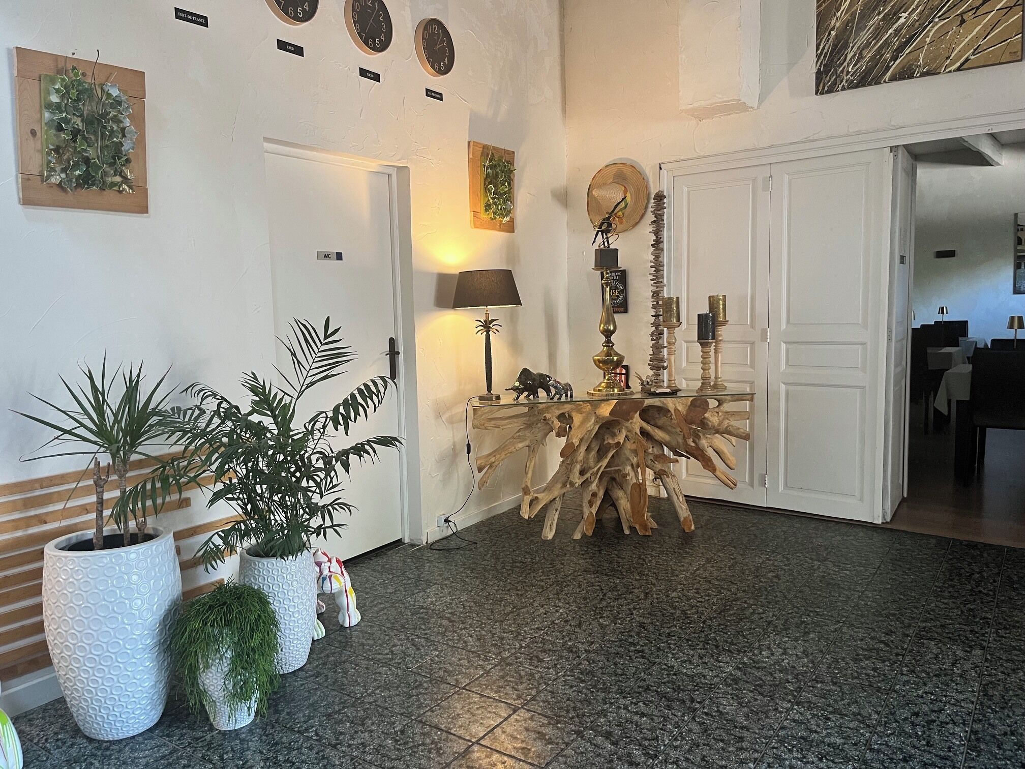 Un hall d’entrée avec un plancher en bois, une table centrale en bois, des plantes en pot, une horloge murale et une porte.
