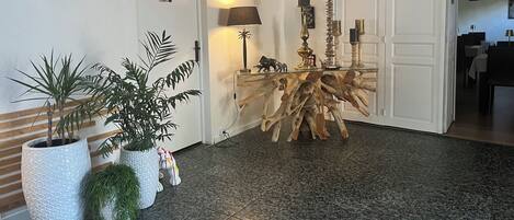 A lobby with a wooden floor, a central wooden table, potted plants, a wall clock, and a door.