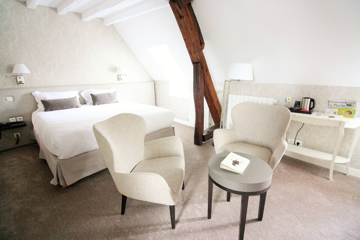 A hotel room with a bed, two armchairs, a small table, and a desk with a lamp and a book.