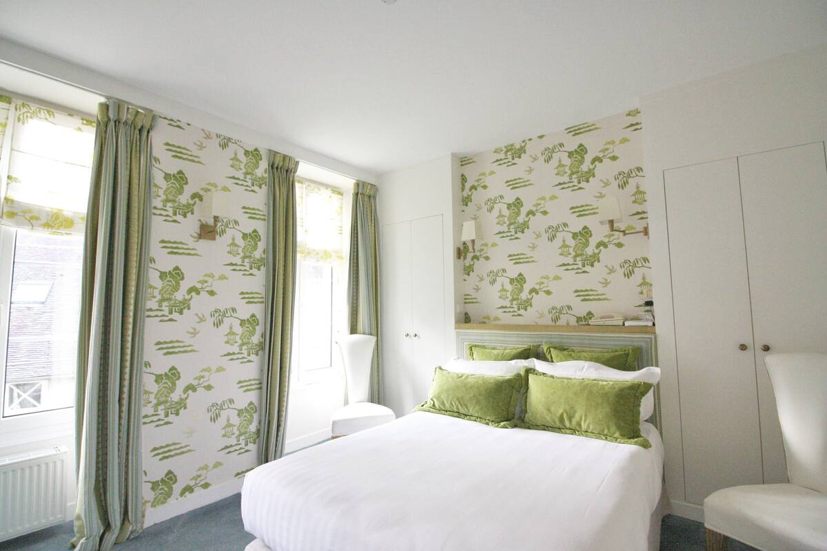 A hotel room with a large bed, green pillows, a white chair, and floral wallpaper.