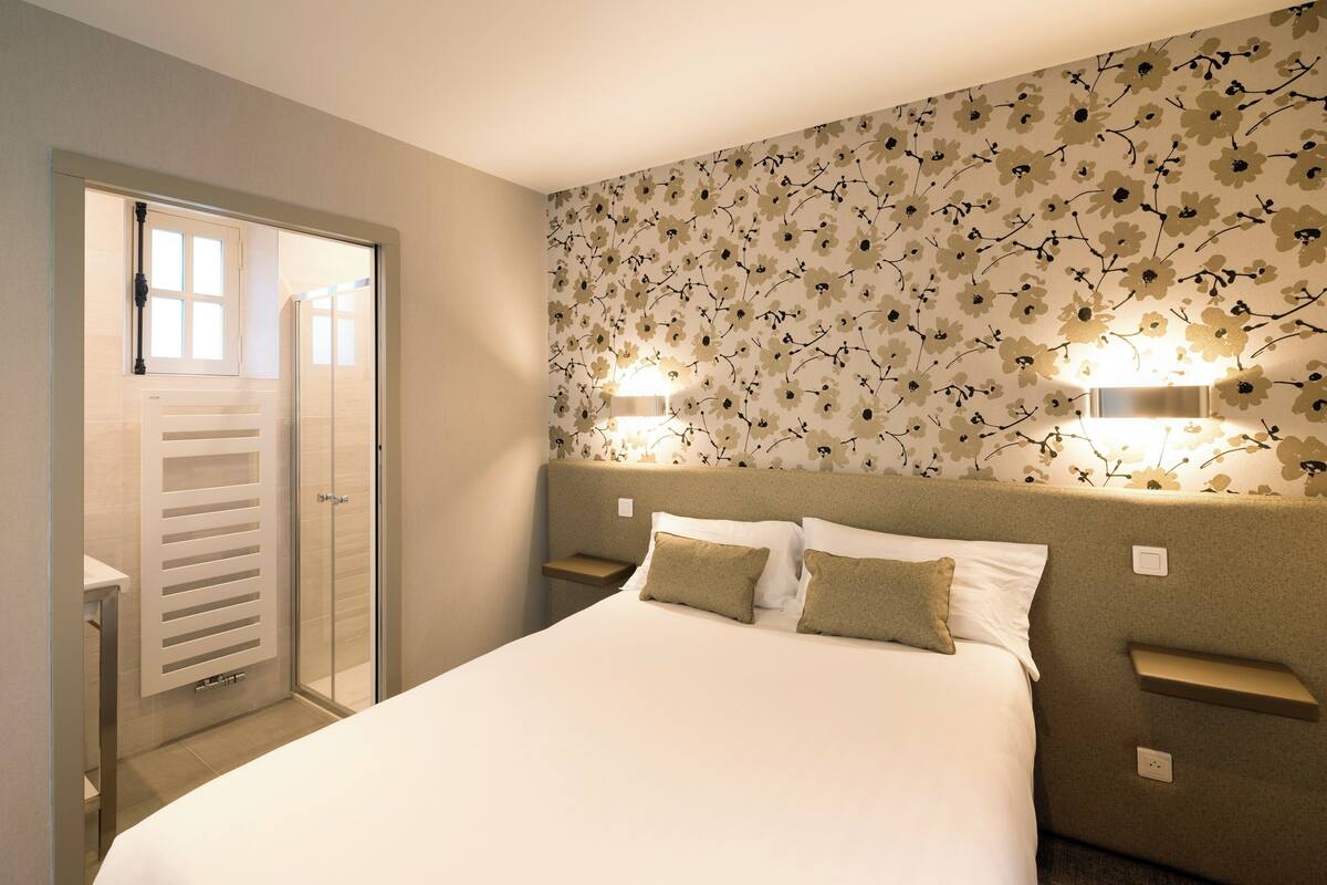 A hotel room with a bed, two bedside tables, a nightstand, a window with blinds, and a wall with a floral pattern.