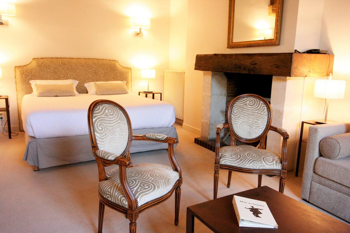 A hotel room with a large bed, two upholstered chairs, a small table, and a fireplace.