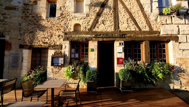 A stone building with a sign reading "Hotel du Tribunal." There are outdoor tables and chairs, and potted plants around the entrance.