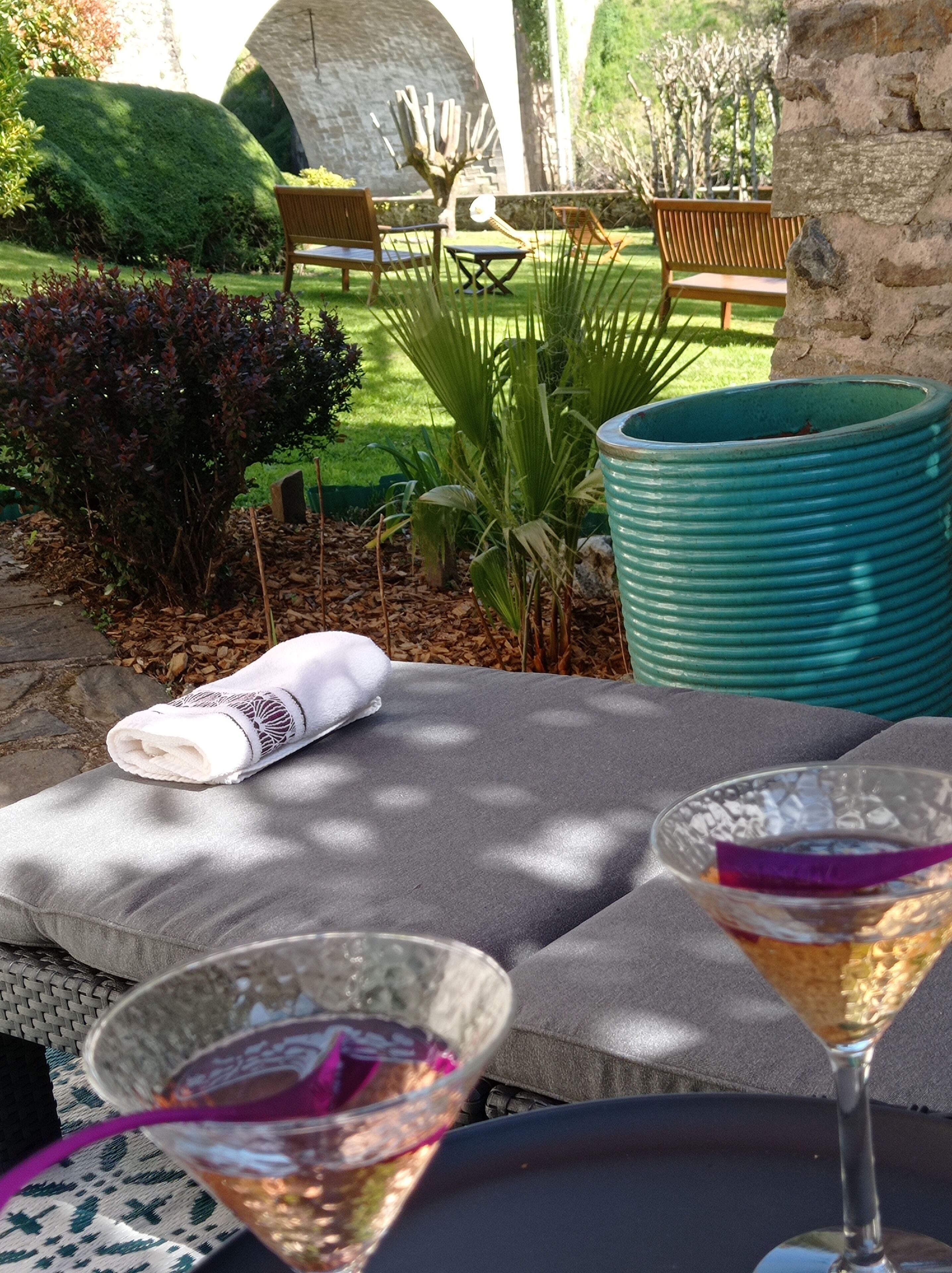 Un jardin avec un mur en pierre, un banc en bois et une table agrémentée d’un coussin et de deux boissons.