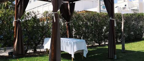 A gazebo with a white tablecloth, surrounded by grass and hedges.