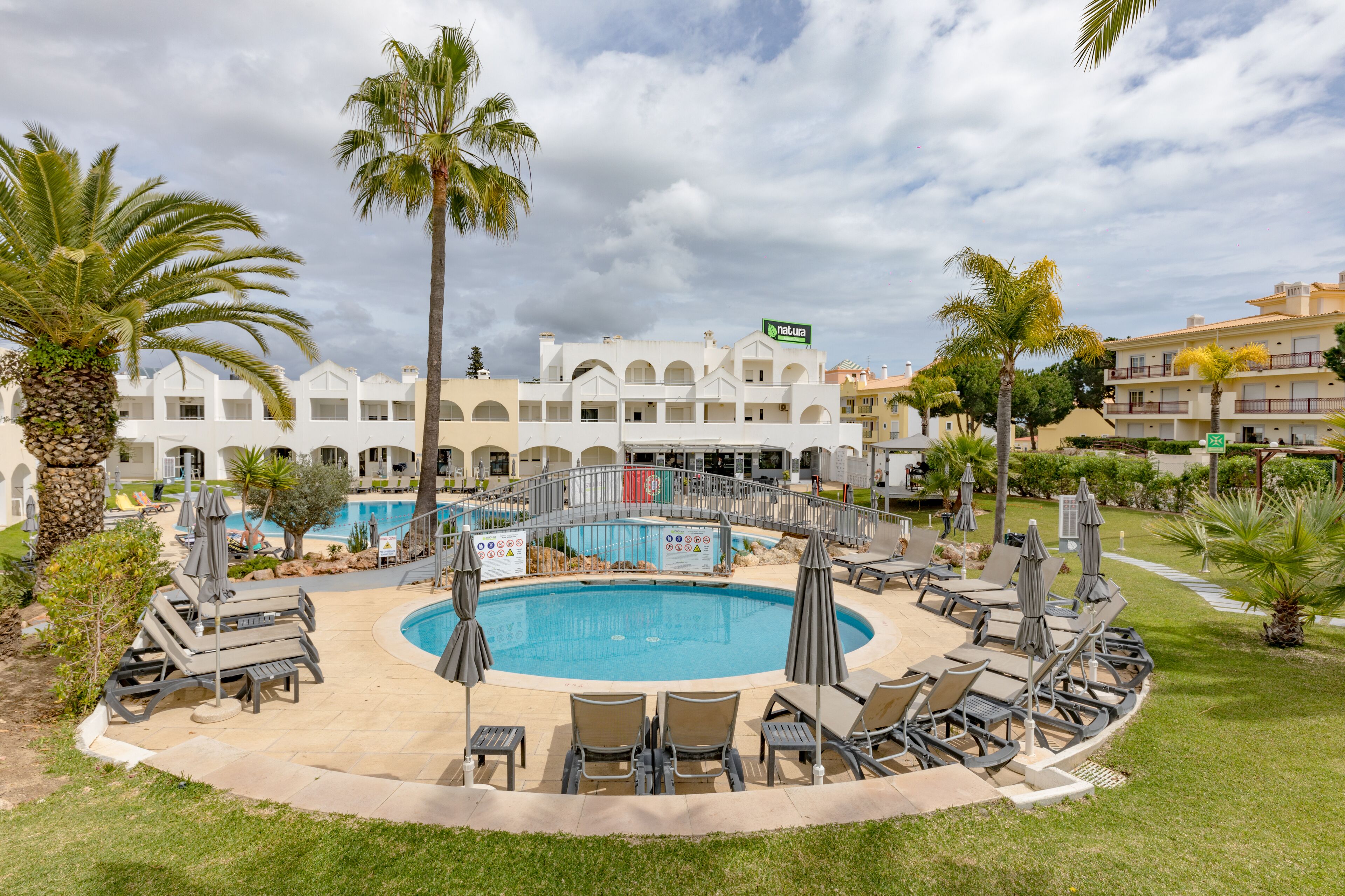 A pool area with lounge chairs and umbrellas, surrounded by palm trees and a building with a sign.