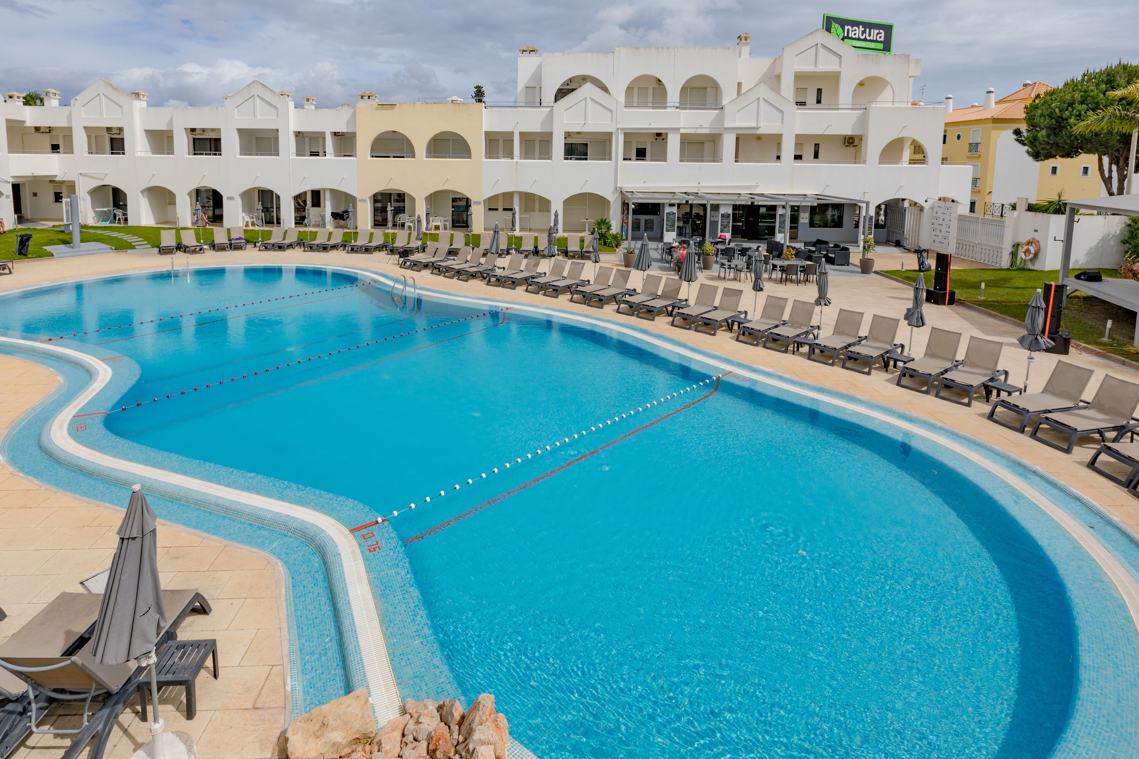 Una gran piscina con varias pistas, rodeada de sillas de descanso y sombrillas, con un edificio al fondo.