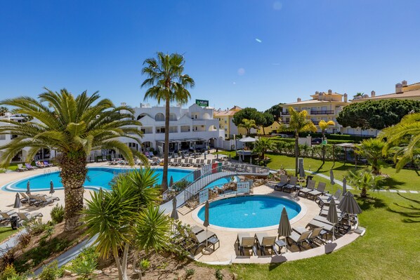 Ein Resort mit zwei großen Schwimmbecken, umgeben von Liegestühlen und Sonnenschirmen, Palmen und blauem Himmel.