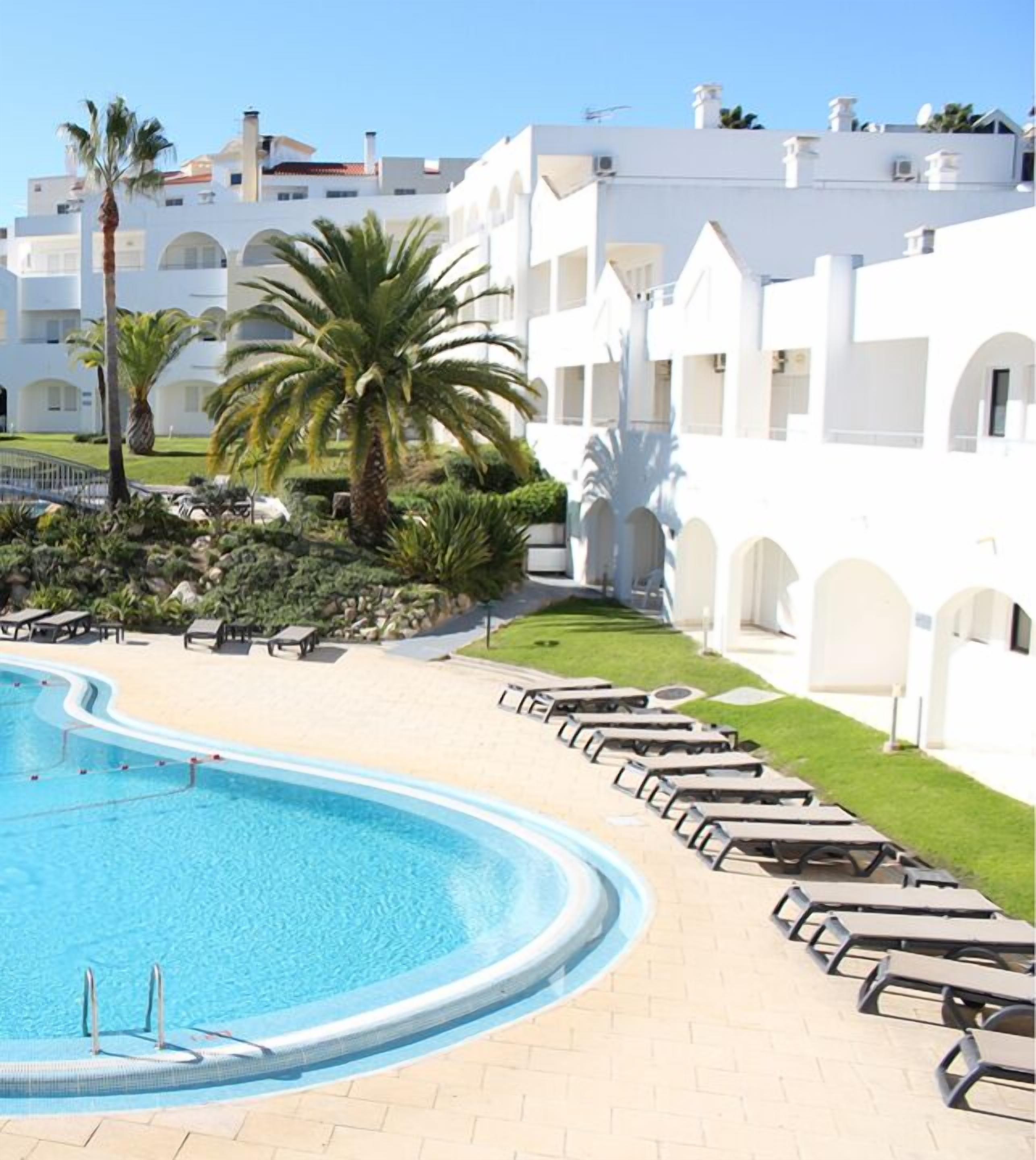 A swimming pool with lounge chairs, palm trees, and a white building with arches in the background.