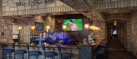 Un bar con pared de piedra, taburetes y una televisión mostrando un partido deportivo.