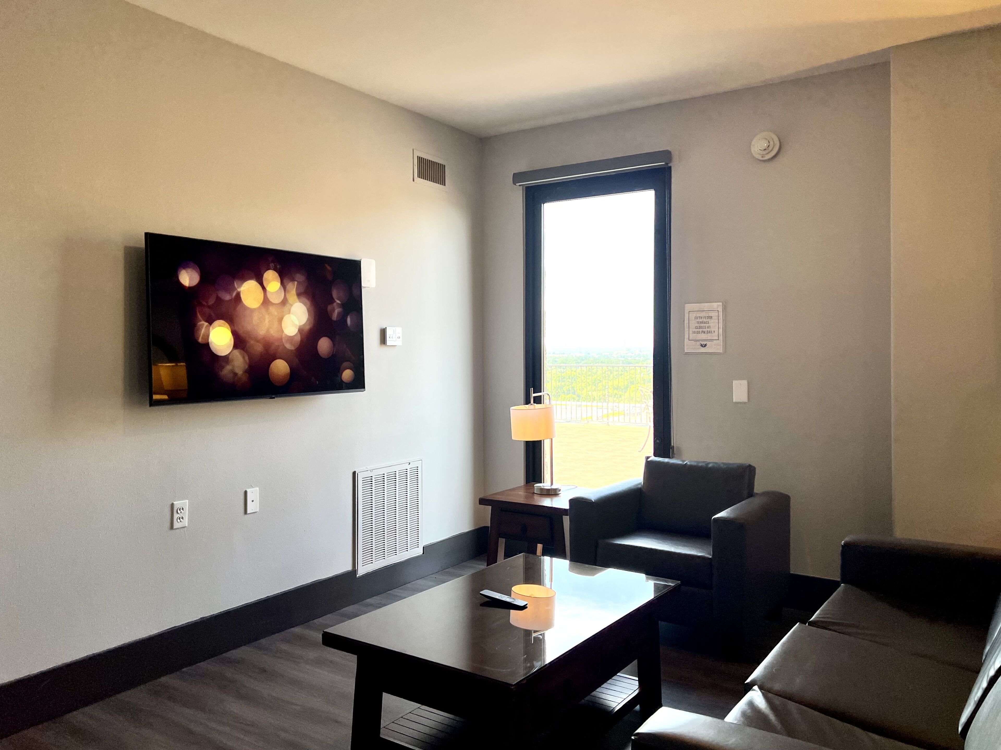 A hotel room with a large window, a flat-screen TV mounted on the wall, a small table with a lamp, and a sofa.