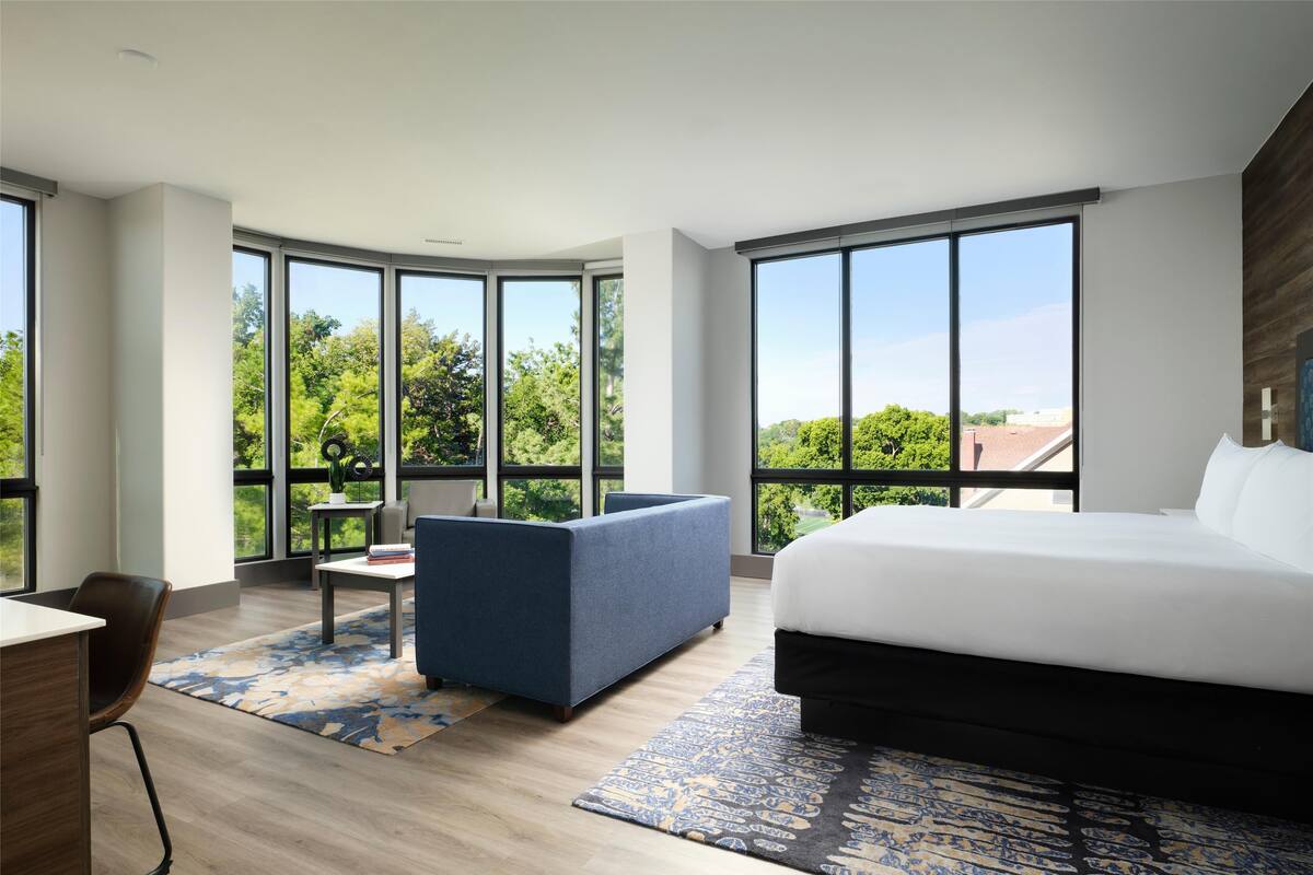 Habitación de hotel moderna con una cama grande, un sofá azul, una mesita pequeña y vistas a zonas verdes a través de amplios ventanales.