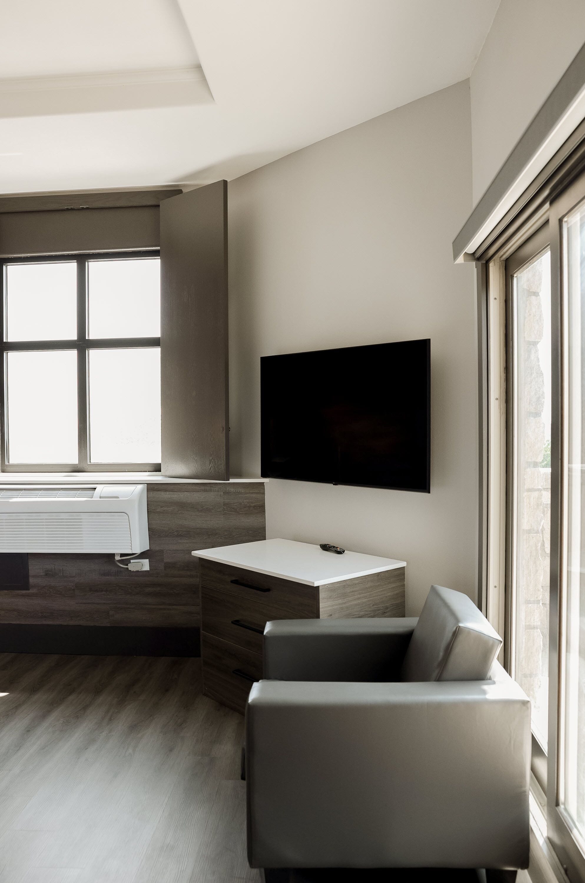 A modern living room with a flat-screen TV mounted on the wall, a built-in air conditioning unit, a grey sofa, and a large window providing natural light.