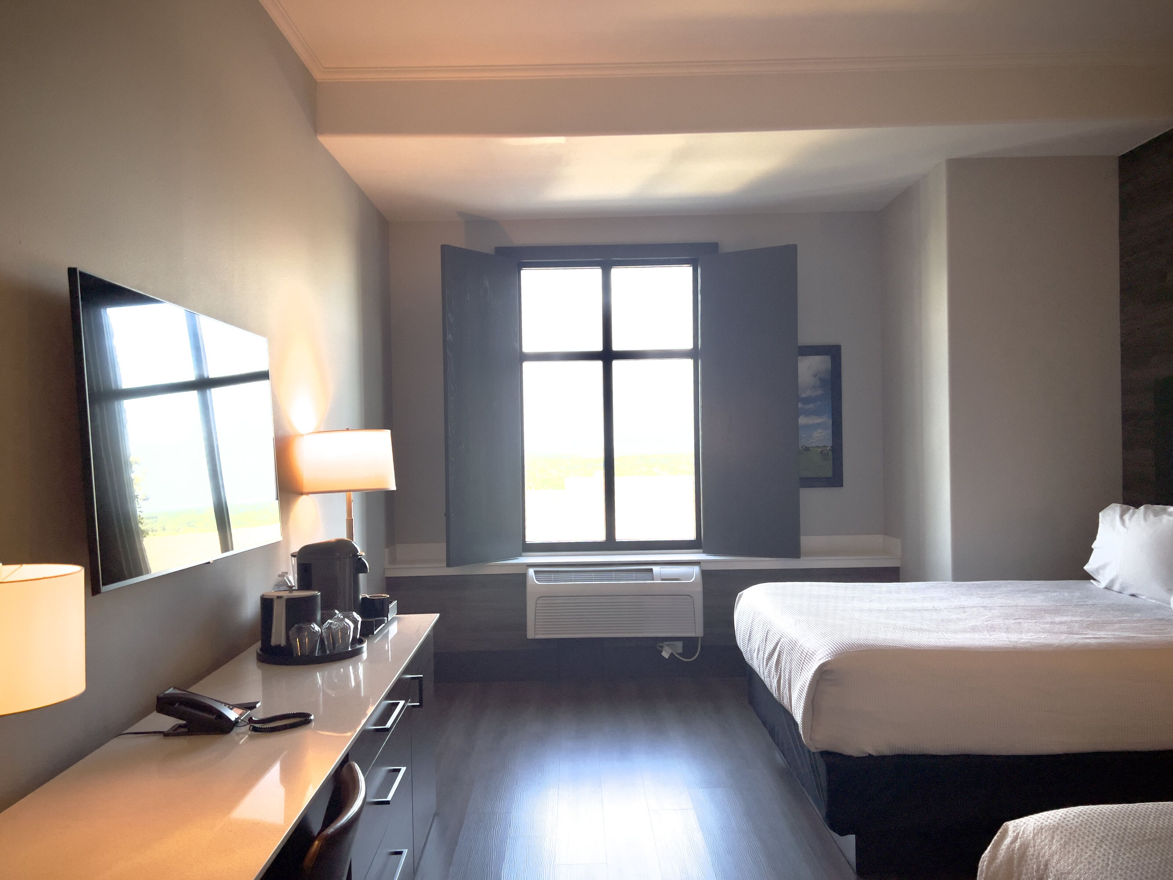 A hotel room with a bed, a desk with a lamp, a telephone, and a window with blinds.