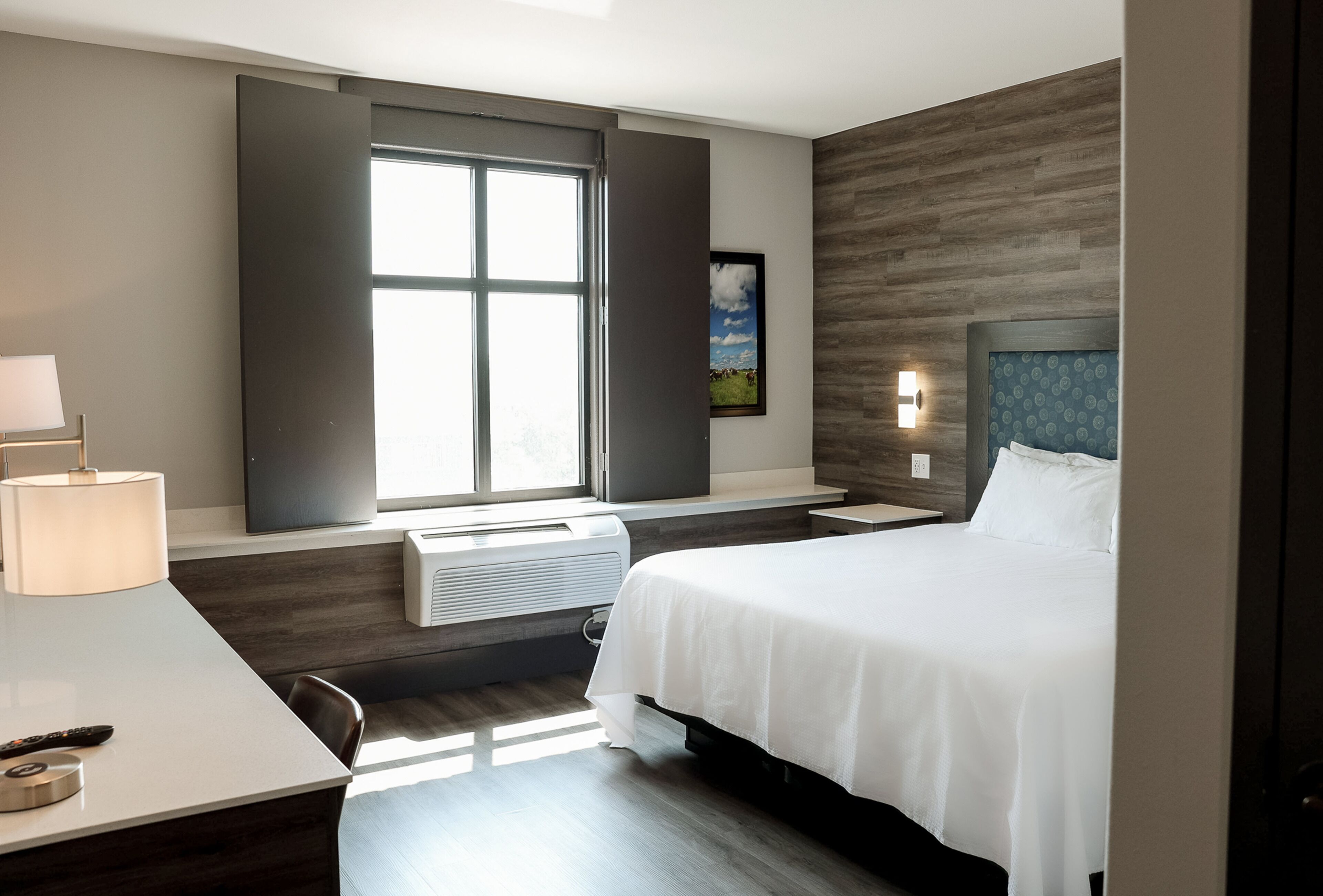 Une chambre d’hôtel moderne avec un grand lit, un bureau avec une lampe, une fenêtre avec des stores, un téléviseur fixé au mur et un tableau encadré.