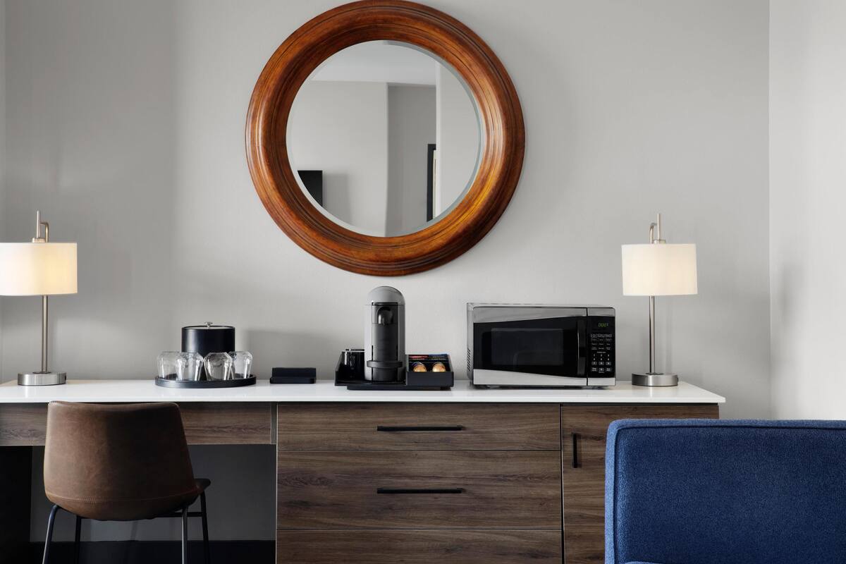 Habitación de hotel moderna con un espejo de marco de madera, cafetera, microondas y dos lámparas sobre una encimera blanca.