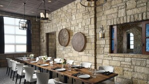 Una larga mesa de comedor dispuesta para una comida en una habitación con paredes de piedra, sillas blancas y lámparas colgantes.