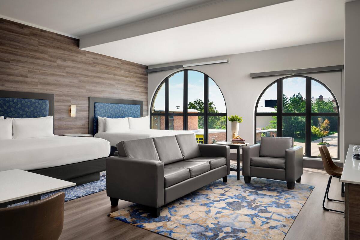 Una habitación de hotel moderna con una cama grande, un sofá gris, una mesita pequeña y dos sillones.