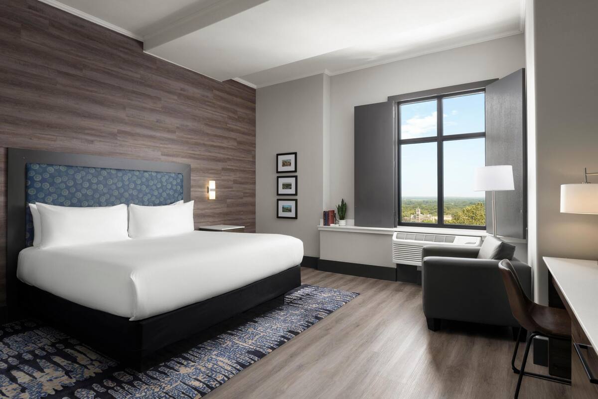 Una habitación de hotel moderna con una cama grande, un escritorio, una silla, un ventanal con vista a la ciudad y un aire acondicionado instalado.