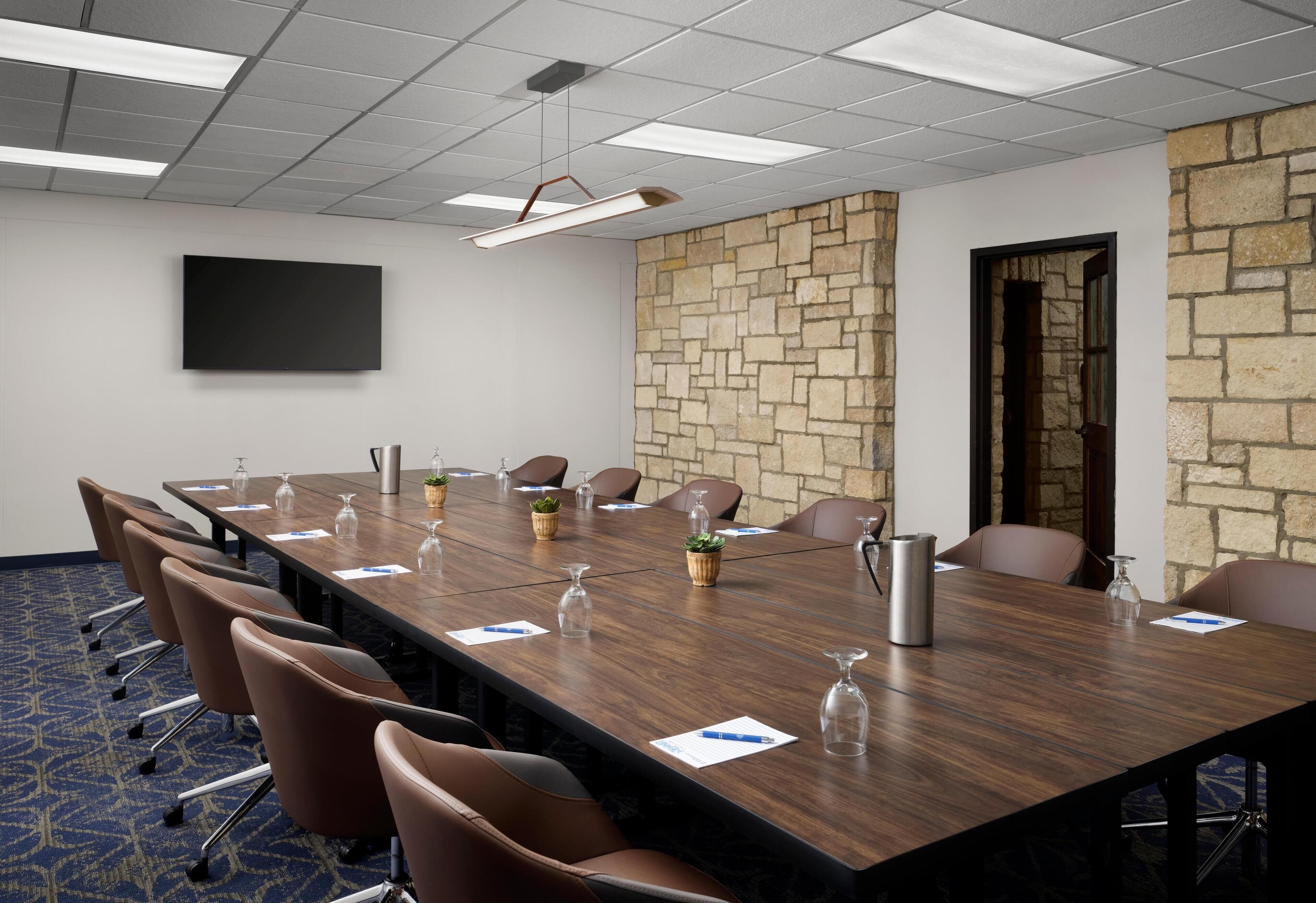 A conference room with a long table, brown chairs, a flat-screen TV, and a stone accent wall.