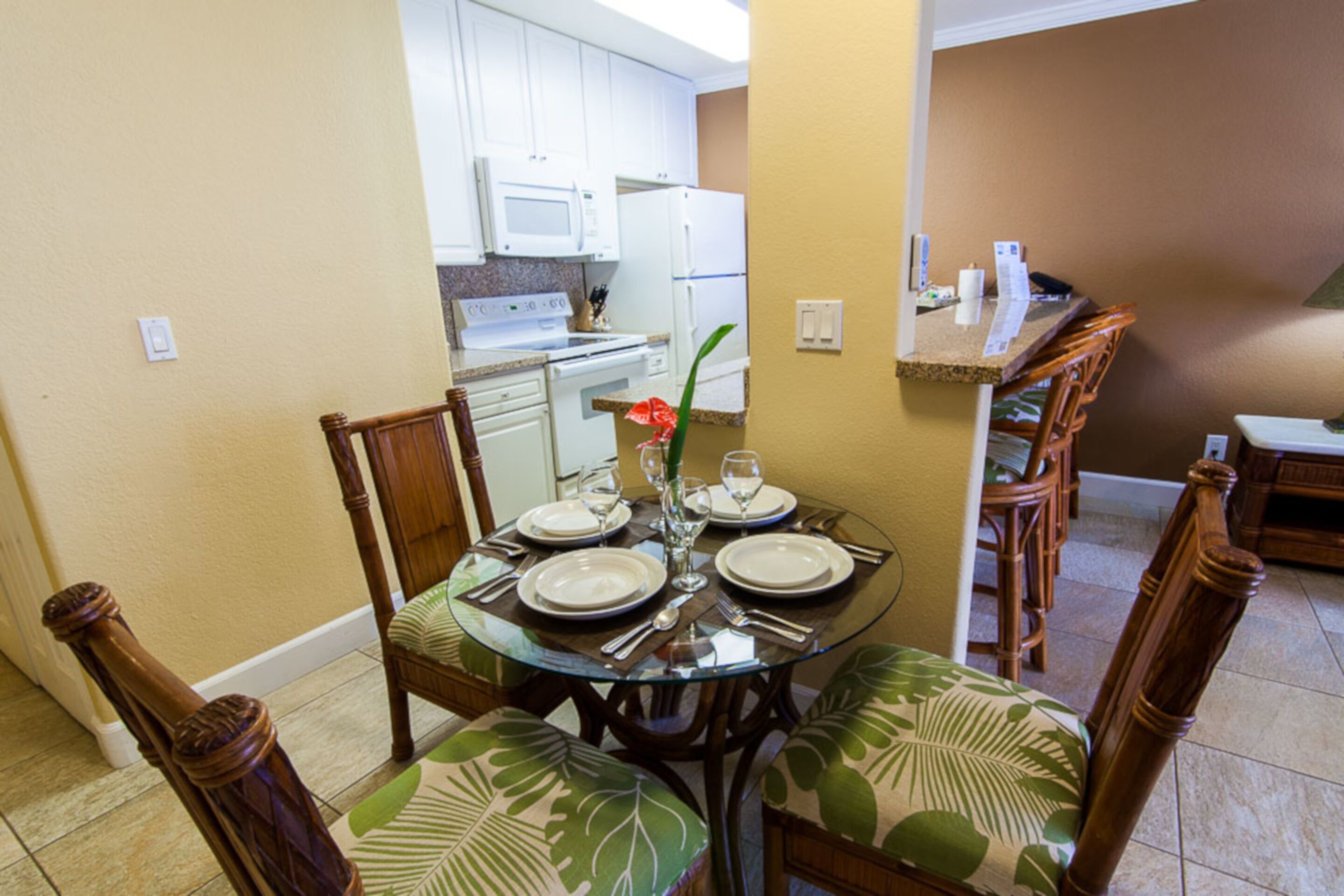 A dining area with a glass table set for four, surrounded by wooden chairs with green leaf-patterned cushions.