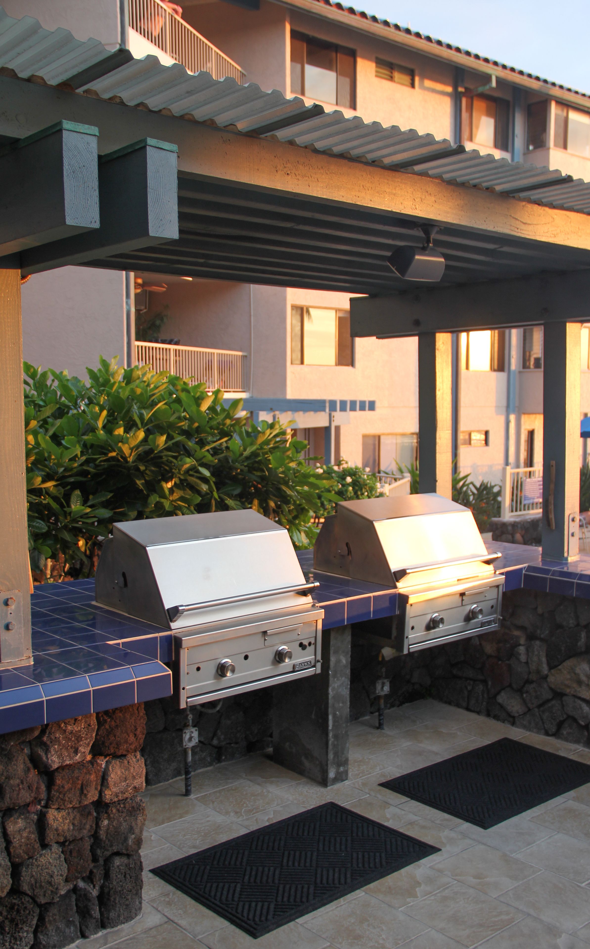 An outdoor barbecue area with two grills, stone pillars, and a covered seating area with a view of the building in the background.