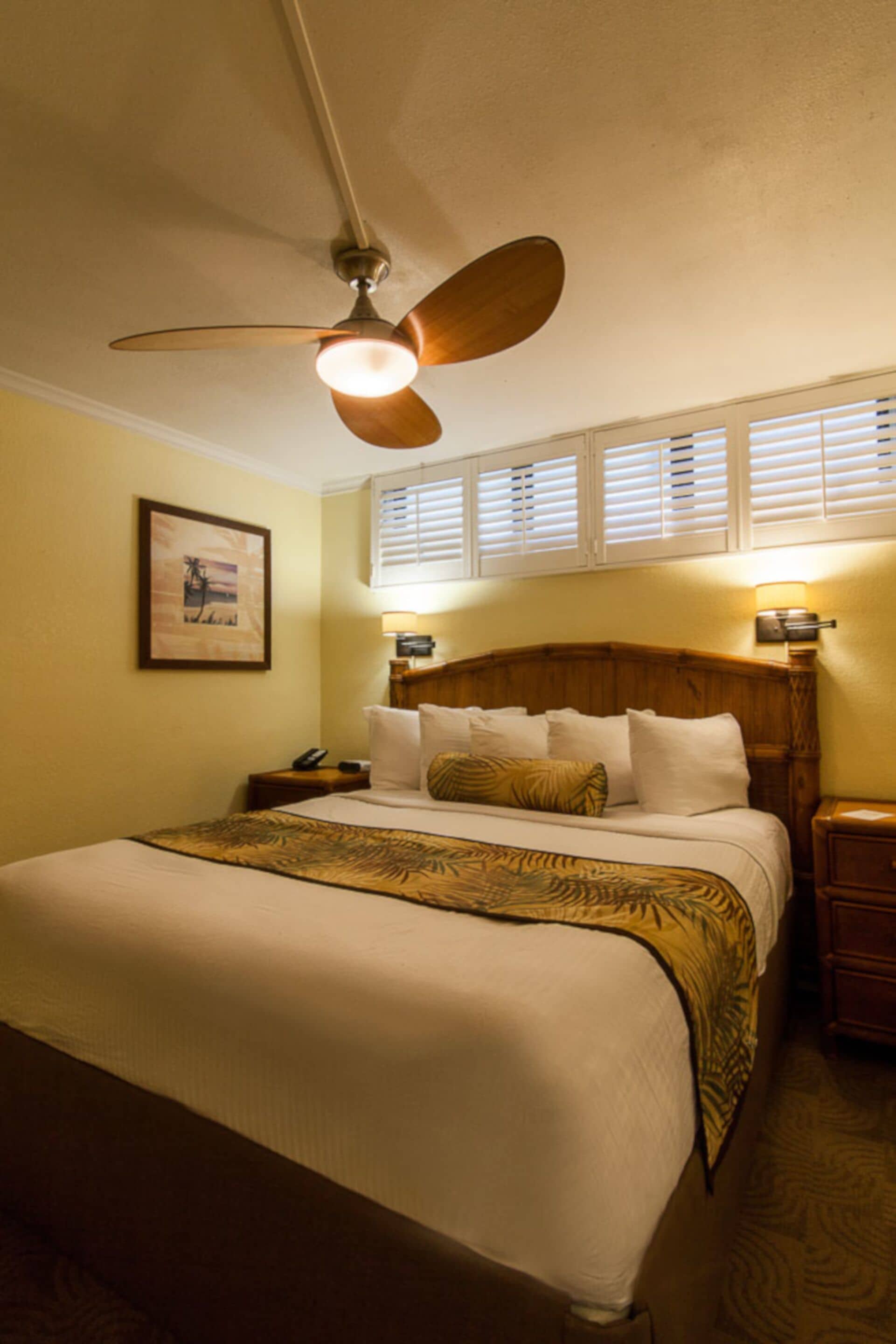 A hotel room with a bed, a ceiling fan, a nightstand, a lamp, and a window with blinds.