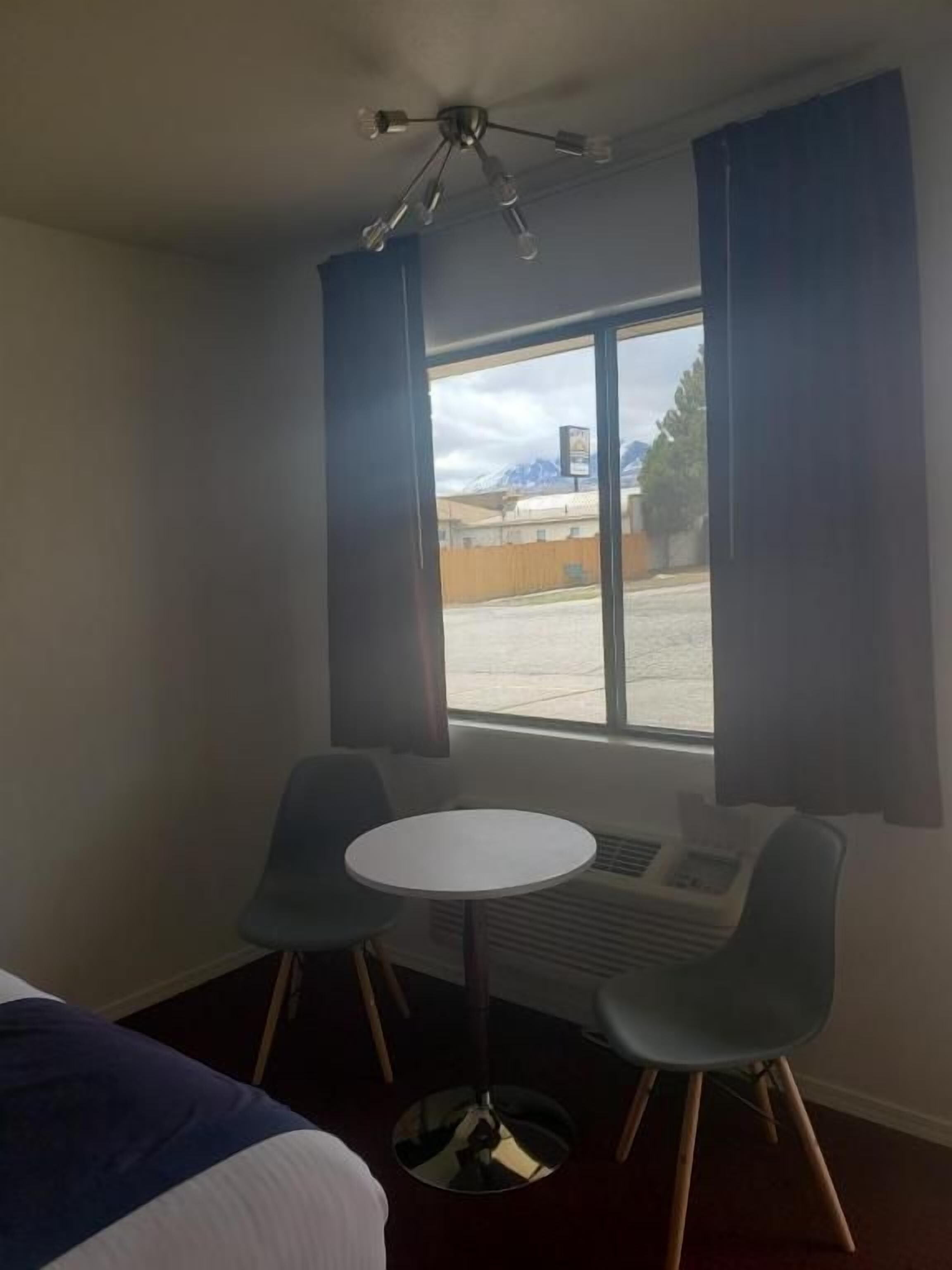 Une chambre d’hôtel avec un lit, une petite table, deux chaises, une fenêtre avec des rideaux et un ventilateur de plafond.