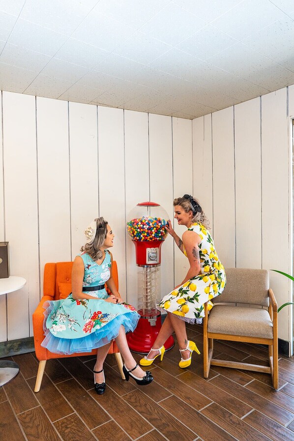 Two women sitting on chairs in a room with wooden panel walls, a red gumball machine, and a small table. - The Atomic Blue Motor-Inn (Monticello)