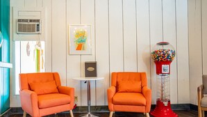 Deux fauteuils orange, une petite table ronde, une œuvre d’art encadrée, une machine à bonbons rouge et un mur blanc avec des panneaux verticaux.